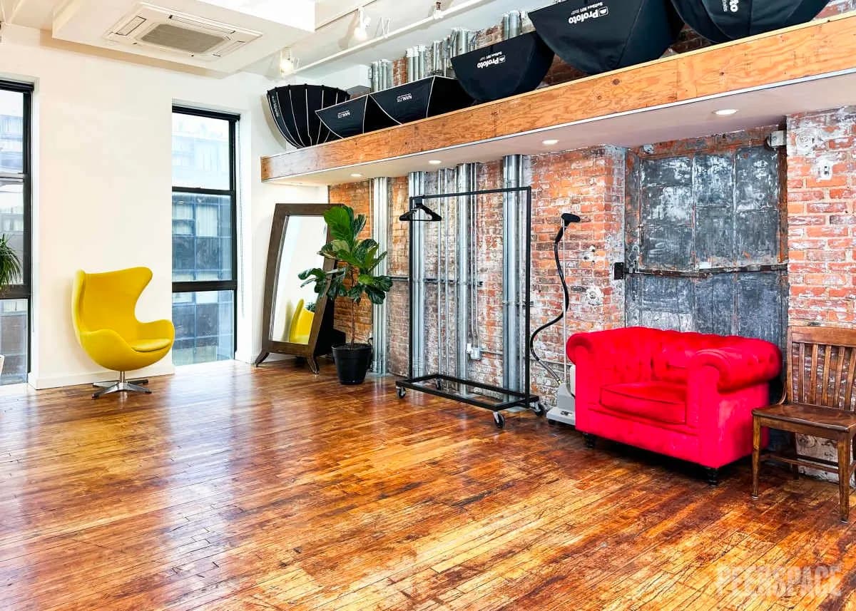 Loft photography studio with a yellow egg chair, red sofa, brick wall, and hardwood floor.