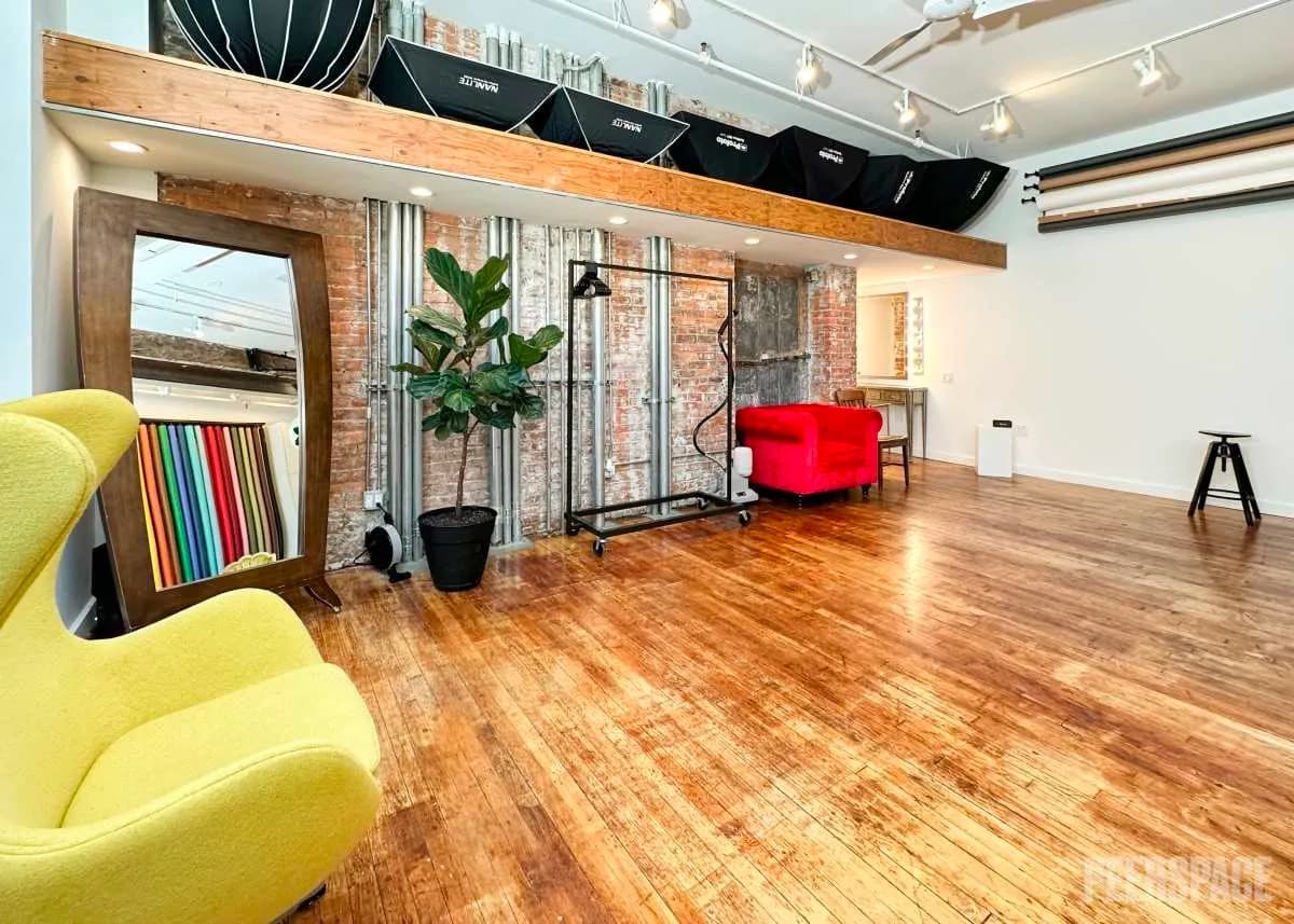 Loft photography studio interior with a yellow egg chair, brick wall, mirror, and hardwood floor.