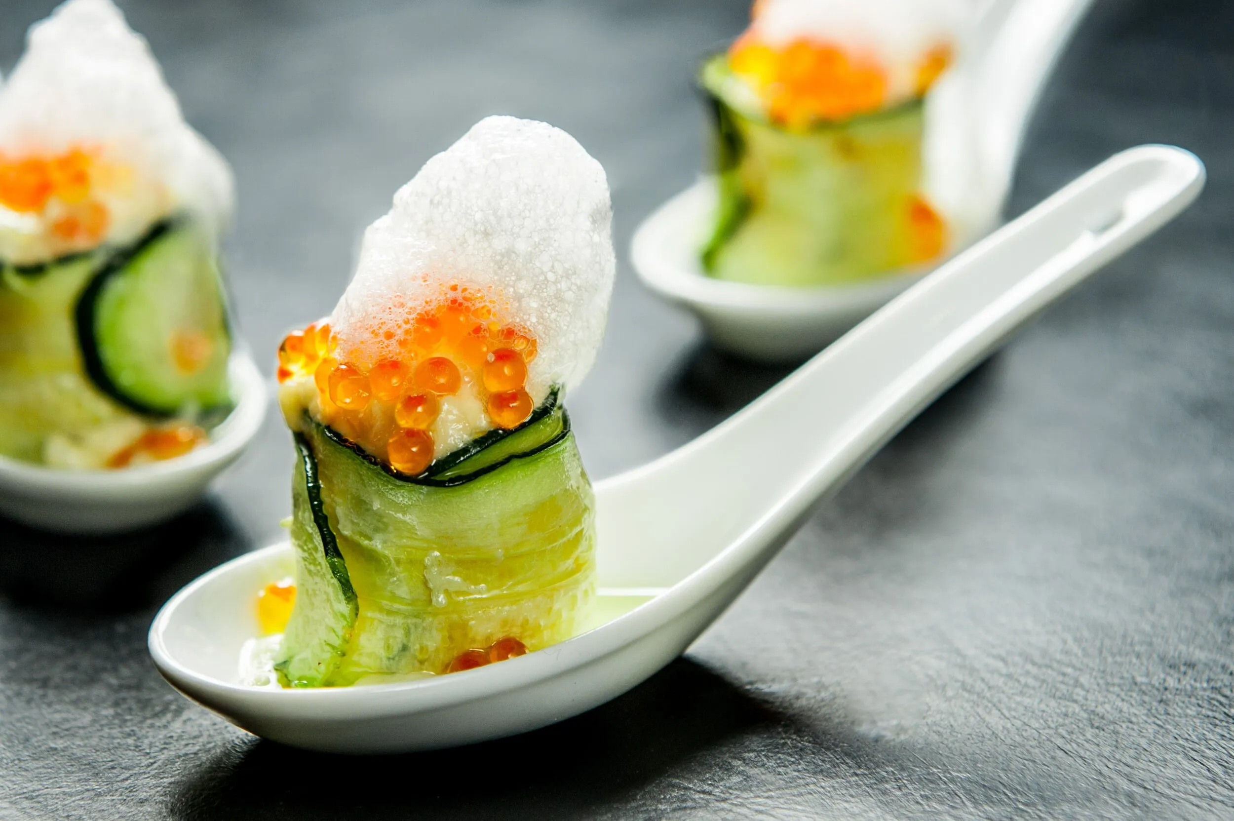 Cucumber-wrapped appetizers topped with orange roe and foam served on white tasting spoons.