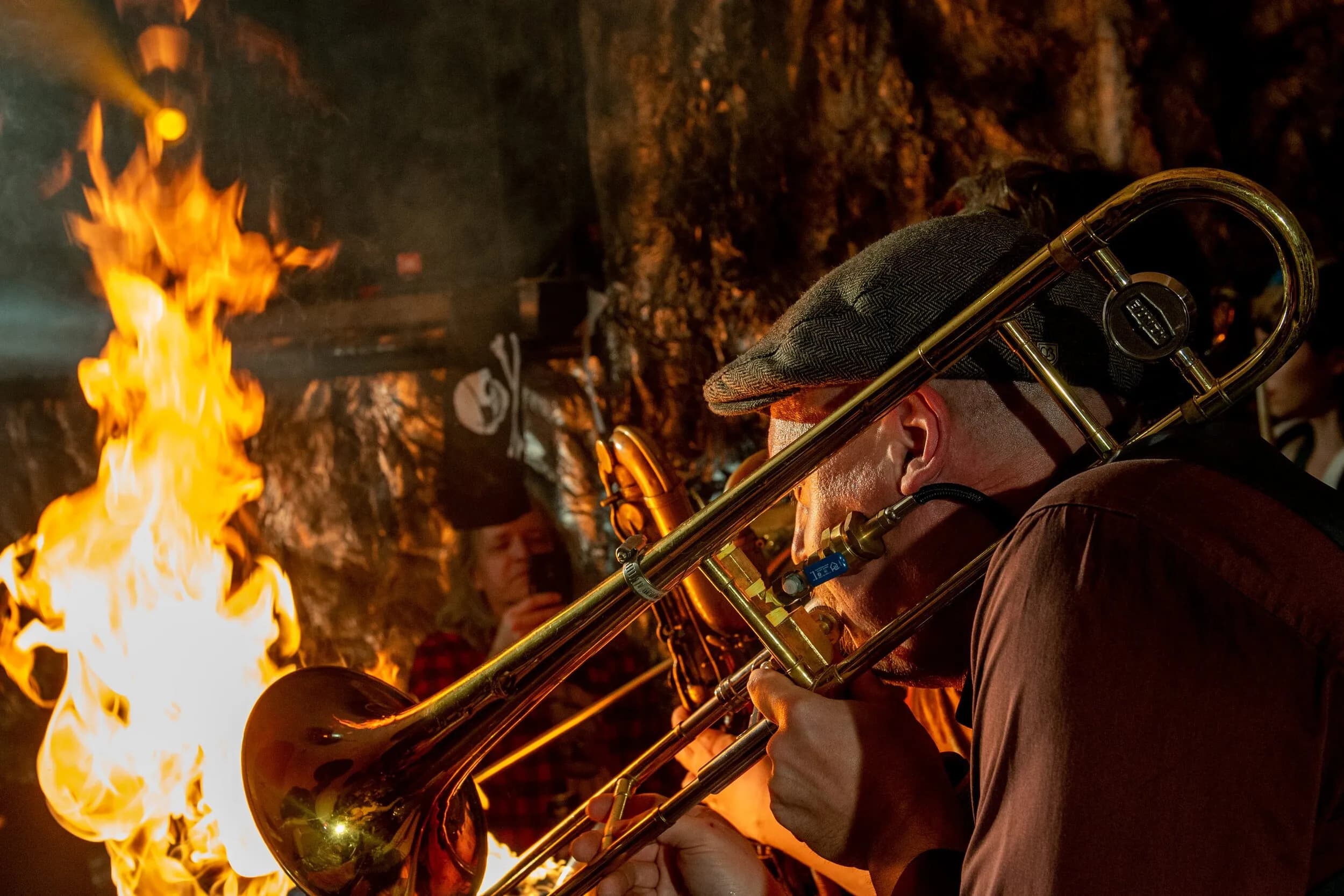 Musician in a flat cap playing trombone next to roaring flames during a fiery live event performance