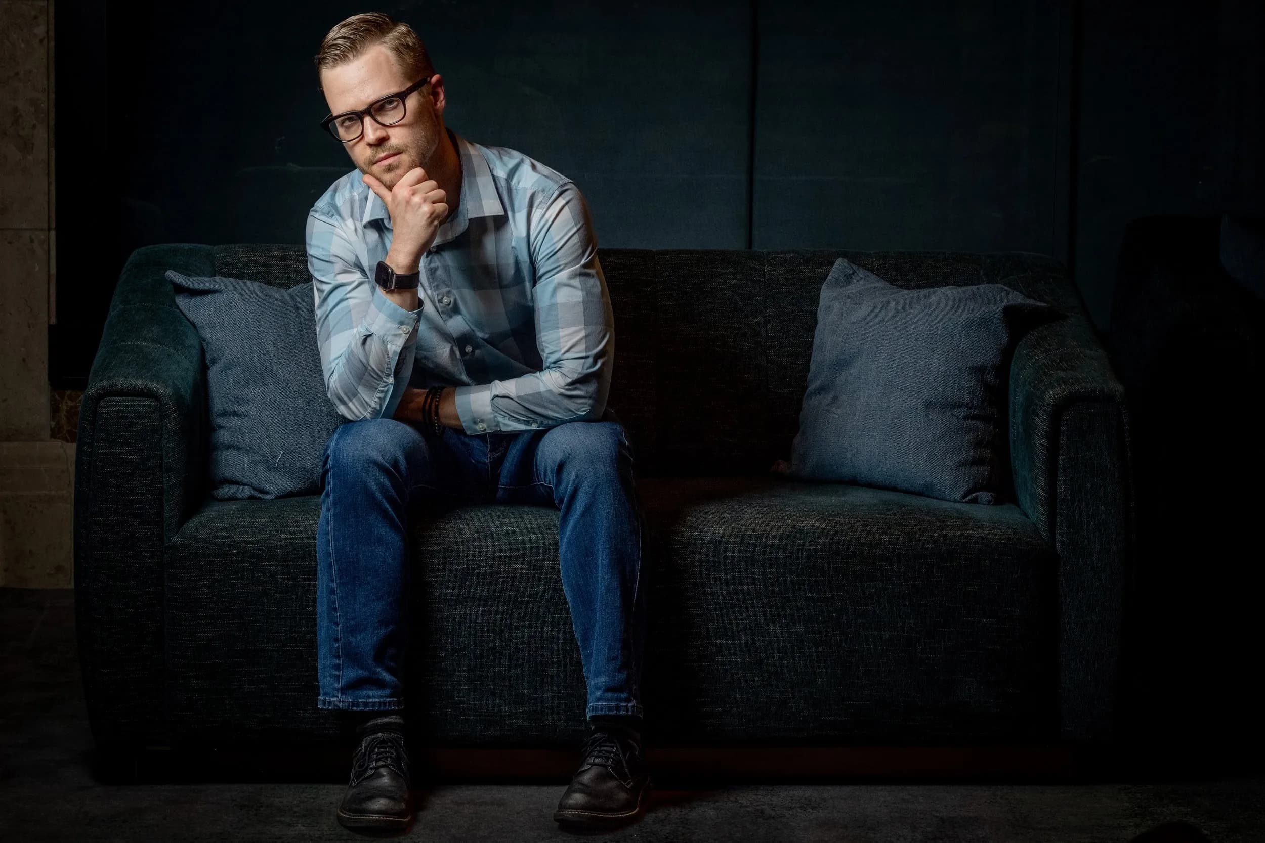 A man in a plaid shirt and jeans sits thoughtfully on a dark sofa with his hand to his chin in dramatic light.