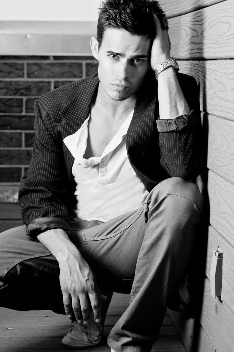 Black and white portrait of a man in a pinstripe blazer and open shirt seated against a brick wall.