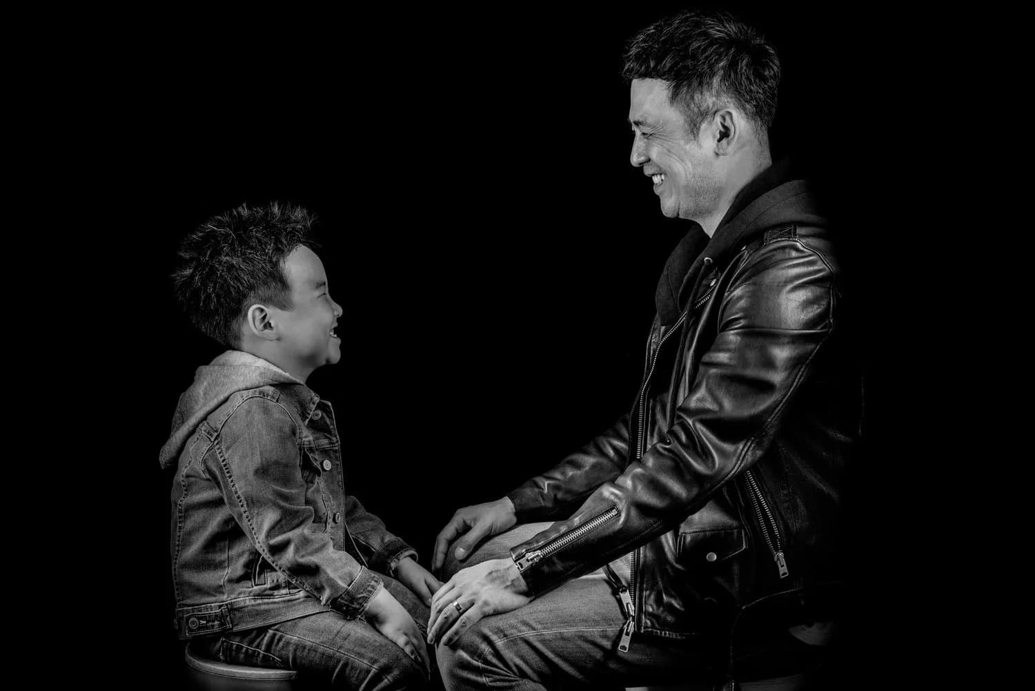 Black and white studio portrait of a man and young boy laughing face to face on a dark background.