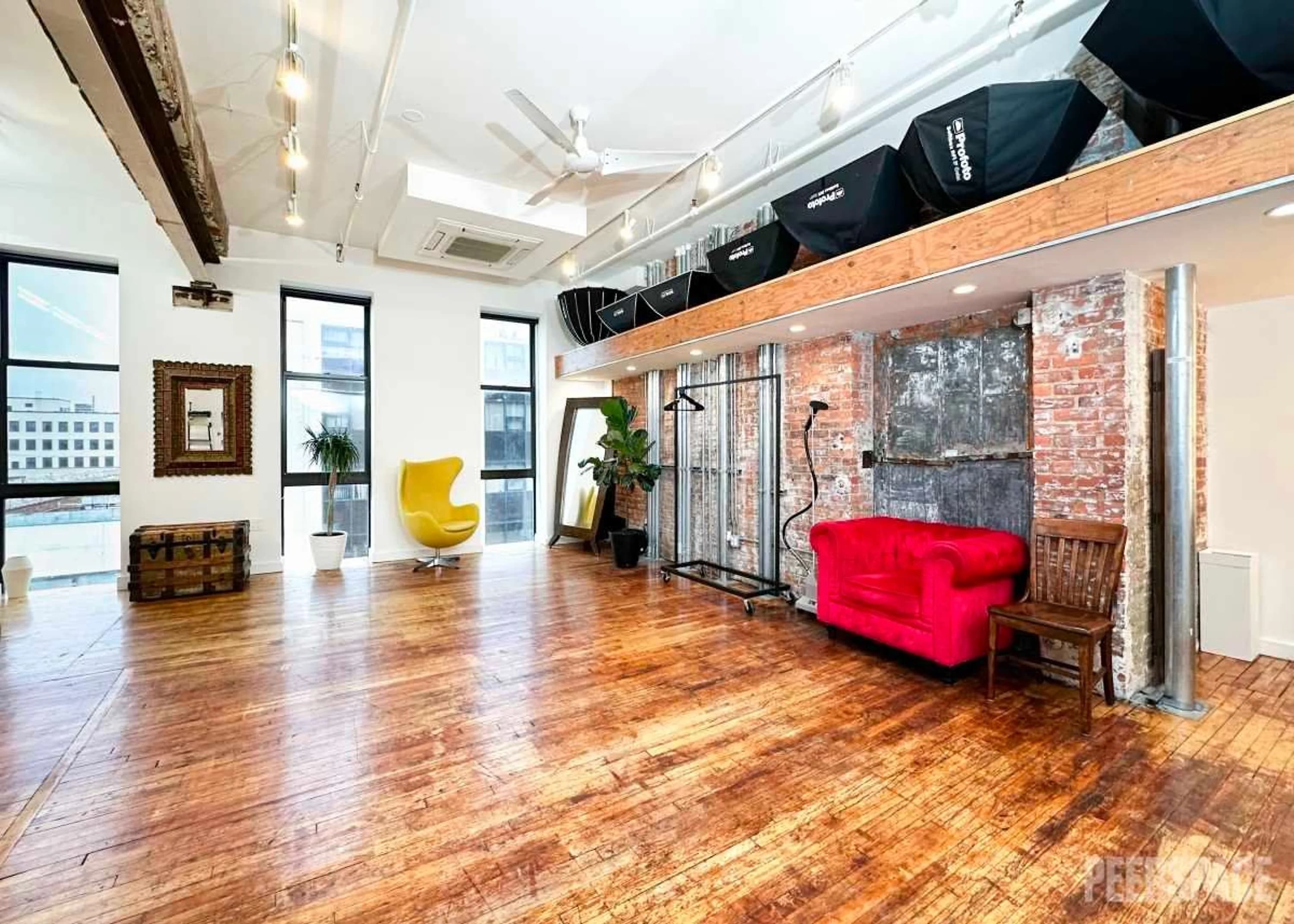 A loft photography studio with reclaimed wood floors, large windows, a red leather sofa, yellow chair and lighting equipment.