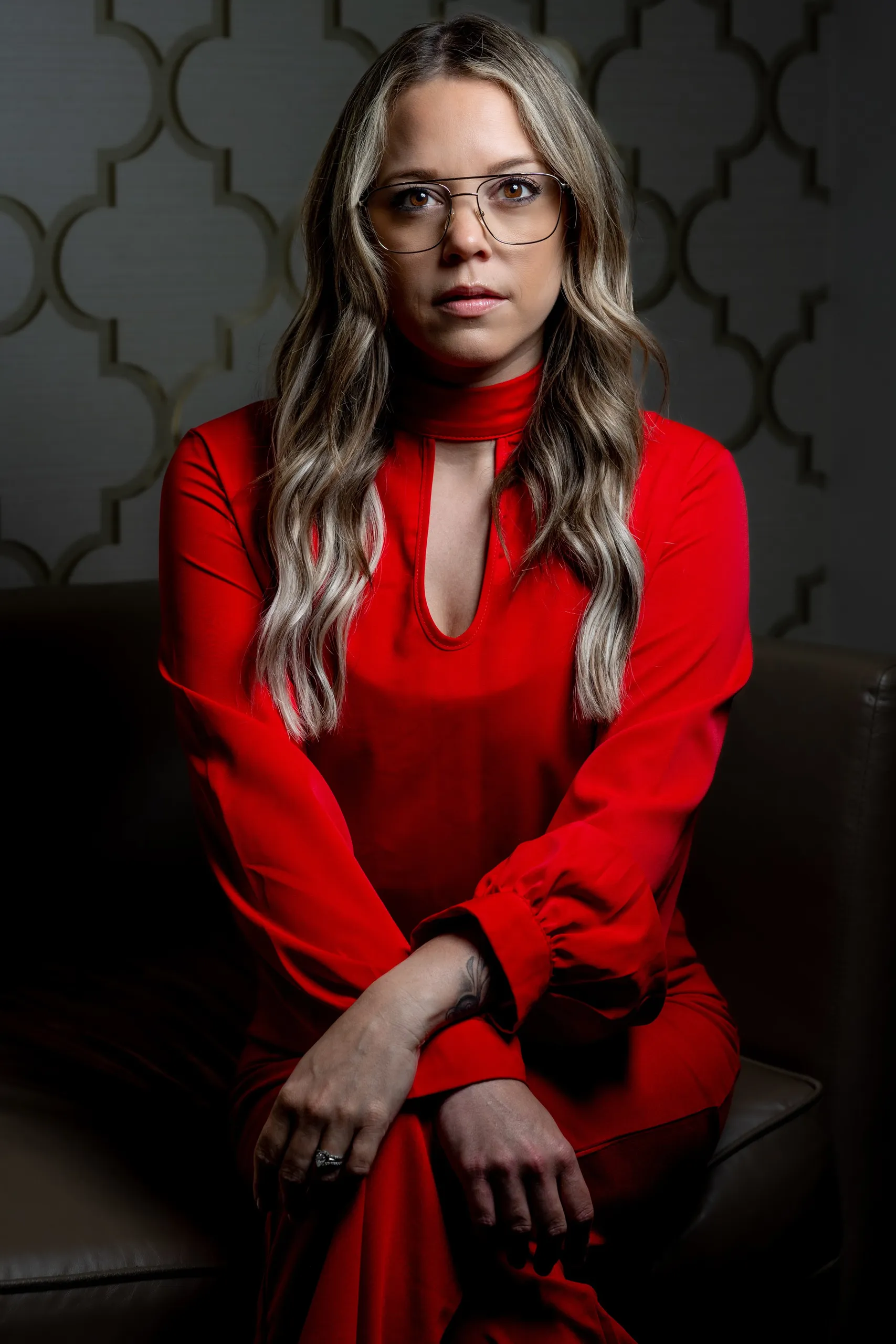 Woman with long wavy hair and glasses wearing a red dress seated in a leather chair against a patterned wall.
