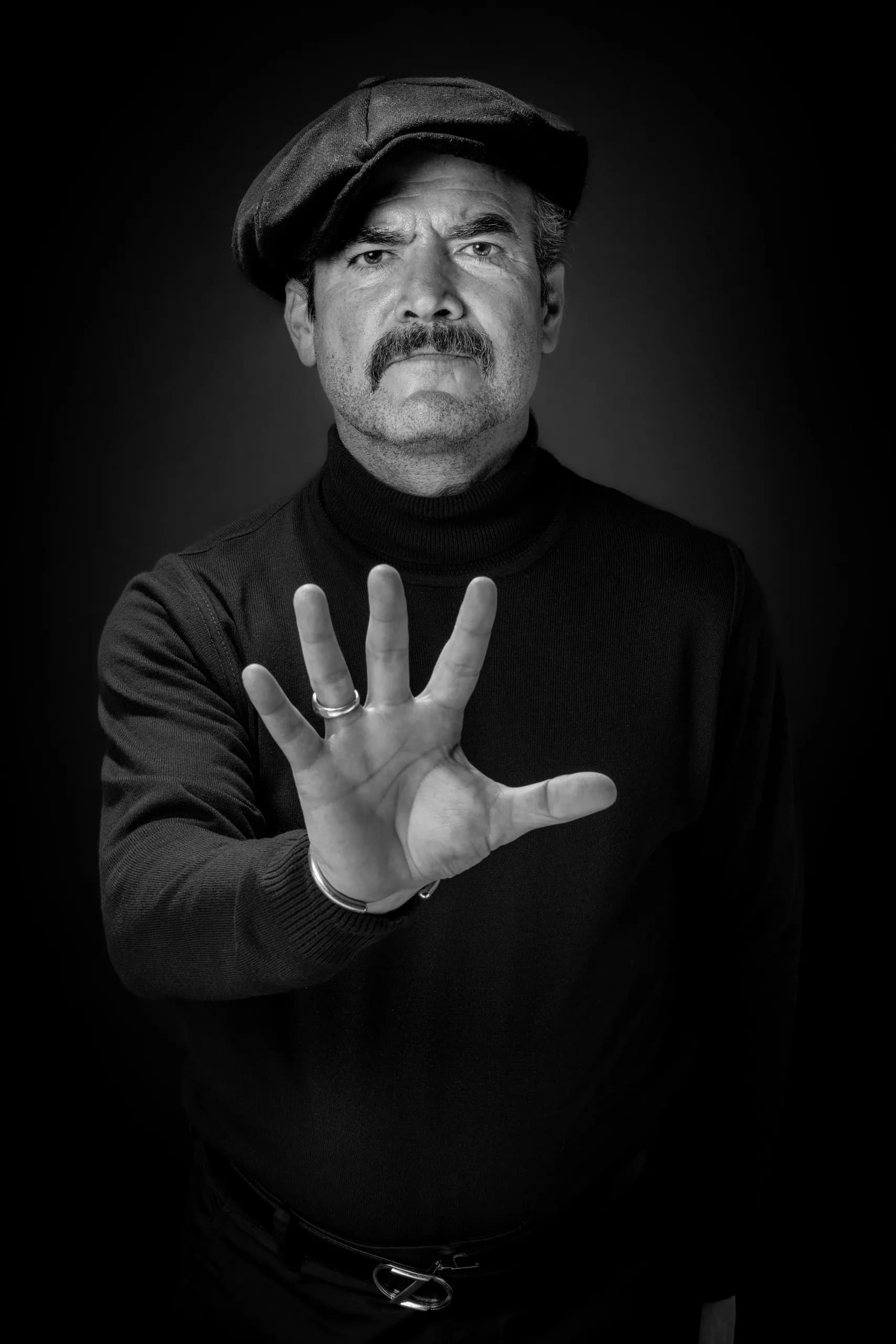 Black and white portrait of a mustached man in a flat cap and black turtleneck holding his open palm toward the camera.