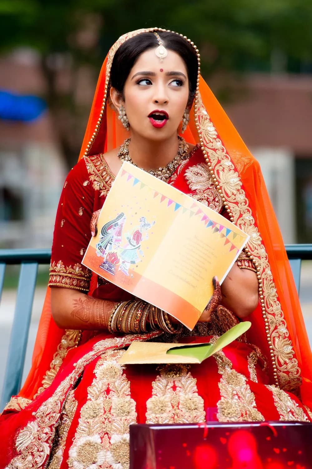 Bride in a red and gold lehenga and orange veil holding an illustrated card while seated outdoors.