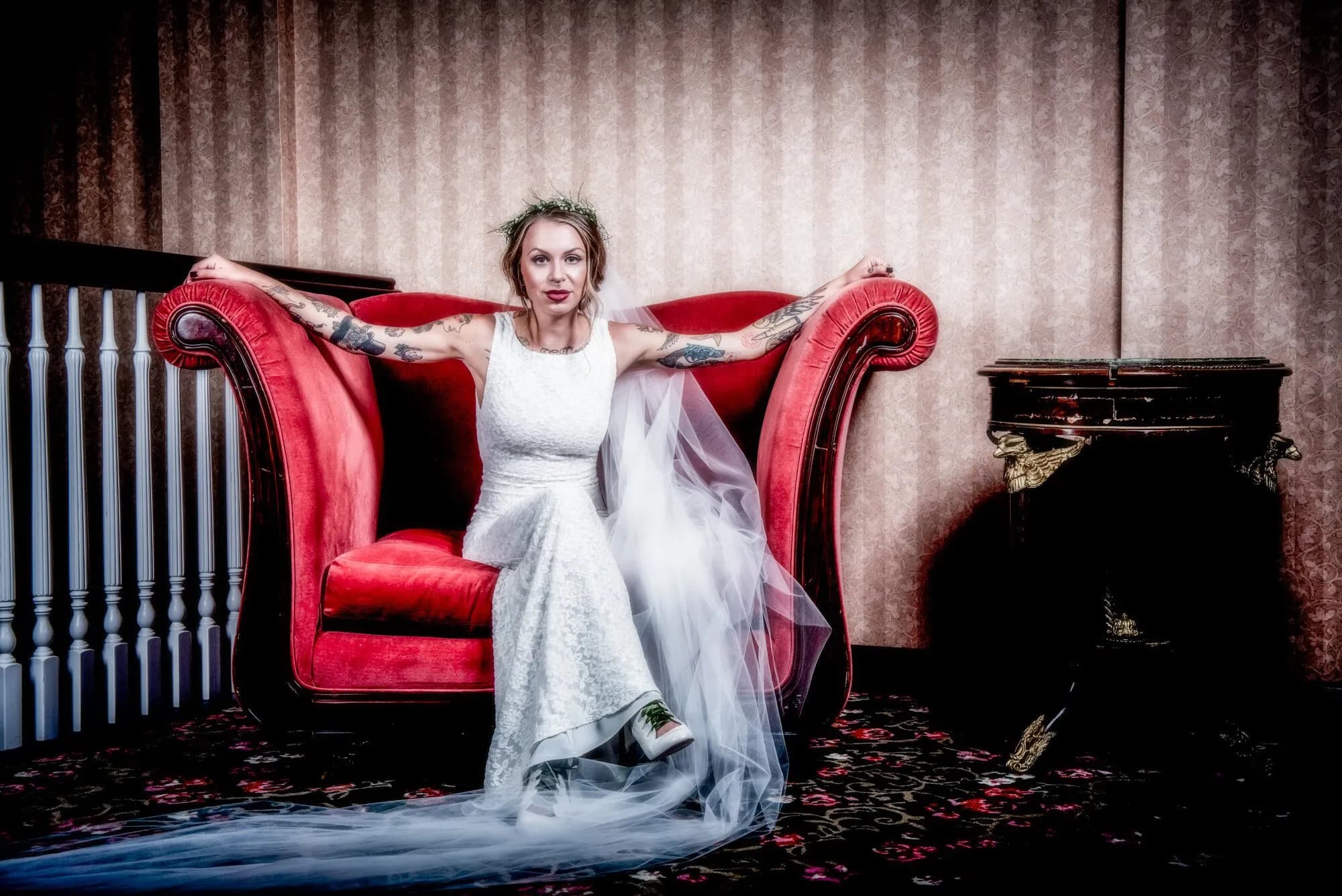 Tattooed bride in a white gown and sneakers reclines on an ornate red velvet sofa with petals scattered on the floor.
