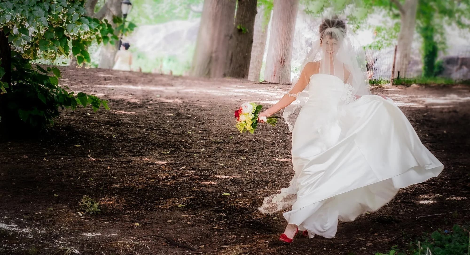 A bride in a flowing white gown and red shoes runs through a wooded park holding a bouquet.